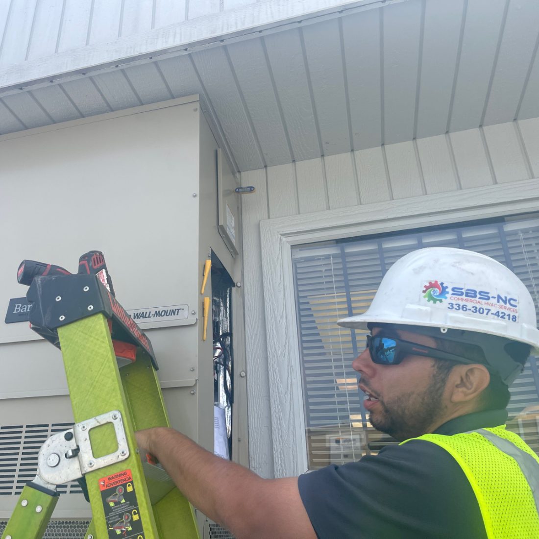 An SBS-NC technician in a hard hat opening the control panel of a wall-mount HVAC unit during a service call